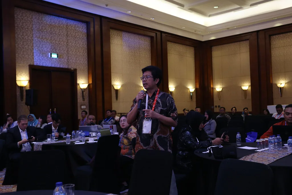 Seorang peserta berdiri mengajukan pertanyaan pada sesi tanya jawab, di antara meja-meja yang masih penuh.